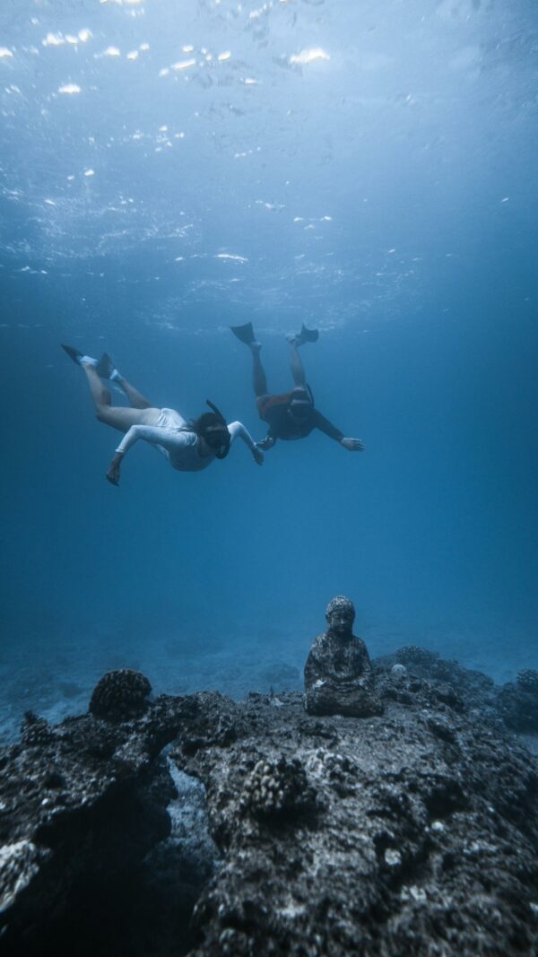 Unrecognizable divers in scuba masks and flippers swimming underwater of blue ocean and observing ancient sunken Buddha statue